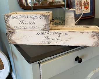 May include: Two white wooden boxes with a distressed finish. The boxes are stacked on top of each other. The top box has the words "French Country Garden" printed on it. The bottom box has the same words printed on it.