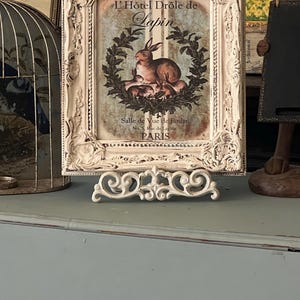 May include: A vintage-style framed print featuring a rabbit illustration with the text "L'Hôtel Drôle de Lapin, Salle de Vue de Jardin, Paris." The frame is off-white with ornate detailing, and it is supported by a decorative stand.