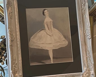 May include: A framed black and white print of a ballerina in a voluminous dress. The print is set in a silver ornate frame and displayed on a metal stand. The ballerina is posed en pointe, with one leg extended.