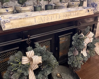 Puede incluir: Una bandeja de madera blanca con las palabras "La Belle Royale" pintadas en ella. La bandeja está llena de pequeños árboles de Navidad en maceta decorados con lazos rojos. La bandeja está sentada en un gabinete negro con dos coronas colgadas de ganchos.