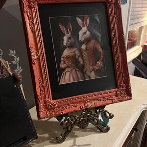May include: A framed print of two white rabbits dressed in formal attire. The frame is a deep red with ornate detailing. The rabbits are standing side by side, the female rabbit on the left is holding a bouquet of flowers. The male rabbit on the right is holding a cane. The print is set on a decorative metal easel.