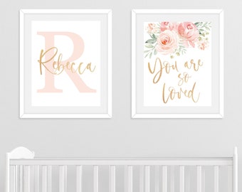 Puede incluir: Dos impresiones enmarcadas para una guardería. La impresión de la izquierda presenta una gran letra R rosa con el nombre Rebecca en escritura dorada. La impresión de la derecha presenta un diseño floral de acuarela con el texto "You are so loved" en escritura dorada.