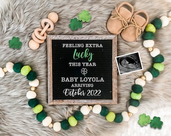 May include: A black letter board with the text "Feeling extra lucky this year" and "Baby Loyola arriving October 2022". The board is surrounded by green and white beads, green shamrocks, and a pair of brown baby shoes.