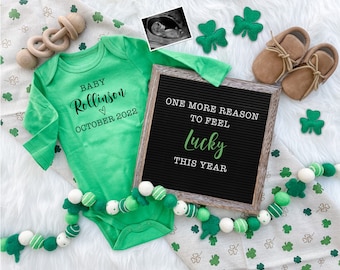 May include: A green baby onesie with the text "BABY Rollinson ♡ OCTOBER 2022". A letter board with the text "ONE MORE REASON TO FEEL Lucky THIS YEAR". A garland of green and white balls with shamrock designs. A pair of brown baby shoes. A white background with green shamrock designs.