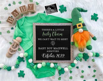 May include: A green and white letter board with the text "There's a little lucky charm. We can't wait to meet Baby Boy Maxwell arriving October 2022". The letter board is surrounded by green shamrocks, a green and white baby onesie, a wooden rattle, and a green and orange gnome.