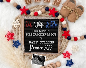 May include: A black letter board with the text "Red, White, & Blue Our Little Firecracker is Due ♡ Baby Collins December 2022". The board is surrounded by red, white, and blue decorations, including stars, beads, and a pair of brown baby shoes.