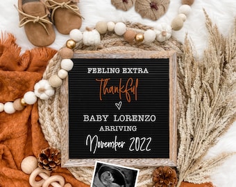 May include: A black letter board with the text "Feeling Extra Thankful" in orange script, "Baby Lorenzo Arriving November 2022" in white text, and a heart symbol. The board is surrounded by a woven straw mat, a garland of white and brown pom-poms, and a small ultrasound image.