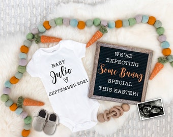 May include: A white onesie with the text "Baby Julio September 2021" and a heart. A letter board with the text "We're expecting some bunny special this Easter!" is next to the onesie. There are carrots and a garland of colorful pom-poms around the onesie and letter board.