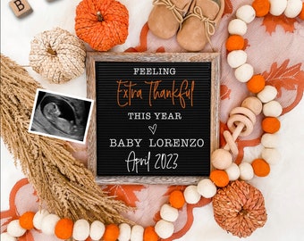 Peut inclure: Un tableau noir avec l'inscription "Feeling extra thankful this year" et "Baby Lorenzo April 2023". Le tableau est entouré de décorations d'automne, notamment des citrouilles, une guirlande de pompons orange et blancs et un hochet en bois.