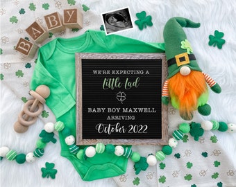 May include: A green baby onesie with a shamrock design, a wooden rattle, a letter block spelling "BABY", a green and white garland, a black letter board with the text "We're expecting a little lad Baby Boy Maxwell Arriving October 2022", a green leprechaun gnome, and a white background with green shamrock patterns.