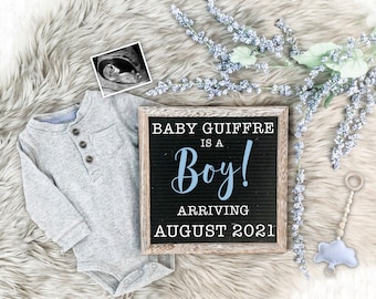 May include: A wooden letter board with the text "Baby Guiffre is a Boy! Arriving August 2021" in white letters. The letter board is on a white faux fur surface with a baby onesie, a sonogram image, and a baby toy.