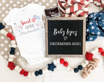 May include: A white onesie with red, white, and blue stars and fireworks, and the text "Sweet Little Firecracker". A black letter board with the text "Baby Lopez" and "DECEMBER 2021". A white, red, and blue felt ball garland with stars.