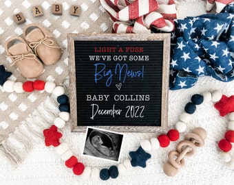 May include: A black letter board with the text "LIGHT A FUSE WE'VE GOT SOME Big News! ♡ BABY COLLINS December 2022". The board is surrounded by red, white, and blue felt balls, a wooden rattle, and a baby ultrasound image.