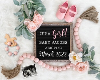 Puede incluir: Un tablero de letras de madera con el texto "It's a Girl! Baby Jacobs Arriving March 2022". El tablero está rodeado de cuentas rosadas, flores y zapatos de bebé.