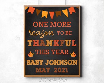 May include: A chalkboard sign with a brown wooden frame announcing a new baby with the text "One more reason to be thankful this year Baby Johnson May 2021". The sign is decorated with autumn-themed decorations, including orange, red, and yellow pennant flags and orange and yellow maple leaves.