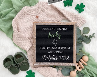 May include: A black letter board with the text "Feeling extra lucky" and a four-leaf clover symbol. Below the text is "Baby Maxwell arriving October 2022". The letter board is surrounded by green shamrocks, a baby sweater, and a wooden rattle.