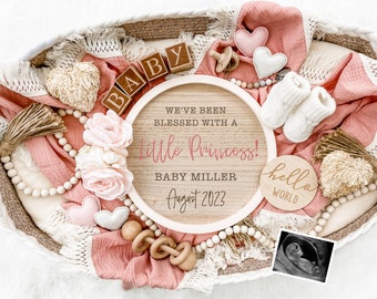 May include: A pink and white baby announcement sign with the text "We've been blessed with a little princess! Baby Miller August 2023". The sign is surrounded by a variety of baby items, including a wooden rattle, a pair of booties, a beaded necklace, and a small wooden sign that says "hello world".