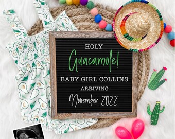 May include: A black letter board with the text "HOLY Guacamole! BABY GIRL COLLINS ARRIVING November 2022". The board is surrounded by a white baby onesie with avocado print, a straw sombrero, a cactus, and a pink maraca.