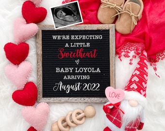 Könnte beinhalten: Eine Buchstabentafel mit der Nachricht "We're expecting a little sweetheart Baby Loyola arriving August 2022". Die Tafel ist umgeben von rosa und roten Filzherzen, einem Paar brauner Babyschuhe, einem roten und weißen Zwerg, der ein rosa Herz mit der Aufschrift "Love" hält, und einer Holzrassel.