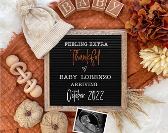 May include: A black letter board with the text "Feeling extra thankful" and "Baby Lorenzo arriving October 2022". The board is surrounded by a brown blanket, a white knitted hat, a wooden rattle, two small pumpkins, and a framed ultrasound image.