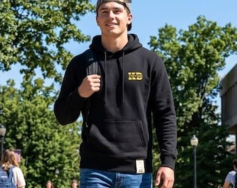 May include: A black hooded sweatshirt with a small yellow logo on the chest, paired with blue denim jeans, a brown baseball cap, and black sneakers. The hoodie features a front kangaroo pocket and a small rectangular tag at the hem.
