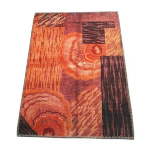 Puede incluir: Una alfombra rectangular con un diseño de patchwork en tonos naranja, rojo y marrón. La alfombra presenta patrones florales y geométricos, con un aspecto texturizado. Los bordes están acabados con un borde gris oscuro.