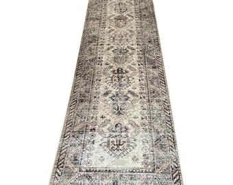 May include: A long, rectangular rug with an intricate, faded pattern. The rug features a central design with geometric shapes and floral motifs in shades of beige, gray, and black. The edges have a darker border with a repeating pattern.