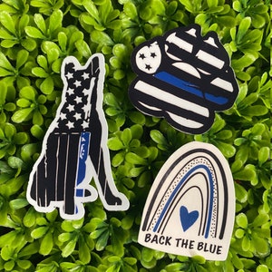 May include: Three stickers with a blue line theme. One sticker is a paw print with a blue line and stars. Another sticker is a dog silhouette with a blue line and stars. The third sticker is a rainbow with a blue line and a heart with the text "Back the Blue".