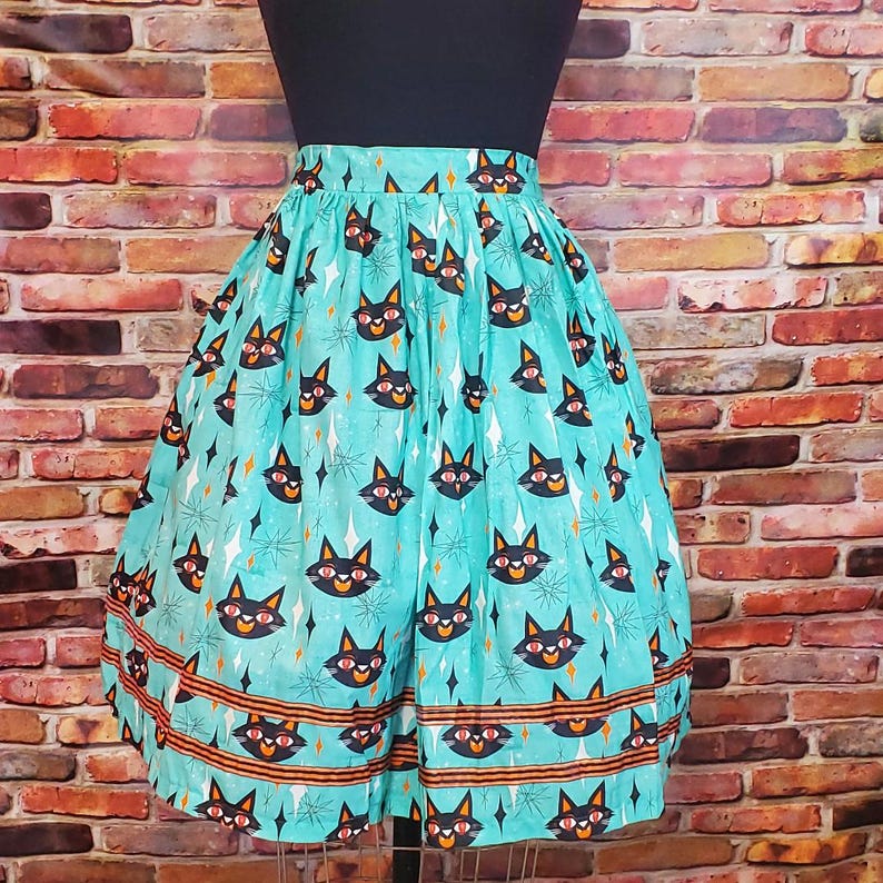 Puede incluir: Una falda azul turquesa con un estampado de caras de gato negro y detalles naranjas. La falda tiene una forma de c&iacute;rculo completo y est&aacute; hecha de una tela ligera.