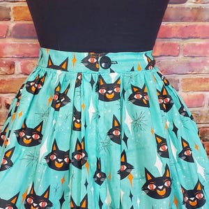 Puede incluir: Una falda azul turquesa con estampado de gato negro y ojos naranjas. La falda tiene una cintura negra y un bot&oacute;n negro.