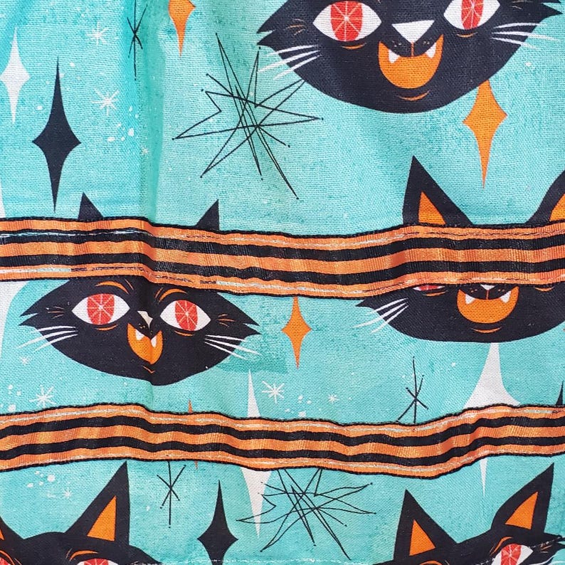 Puede incluir: Una tela turquesa con un patr&oacute;n repetido de gatos negros con ojos naranjas y un patr&oacute;n de rayas negras y naranjas. El fondo tiene un dise&ntilde;o retro de estrella.