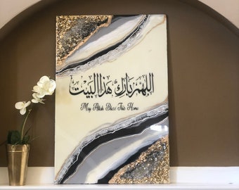 May include: A white canvas with black and gold abstract art. The Arabic text "اللَّهُمَّ بَارِكْ لَنَا فِي هَذَا الْبَيْتِ" is written in black calligraphy. The text "May Allah Bless This Home" is written in black below the Arabic text.
