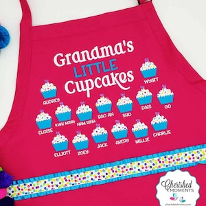 May include: Pink apron with the text "Grandma's Little Cupcakes" and a row of cupcakes with names printed below each one.