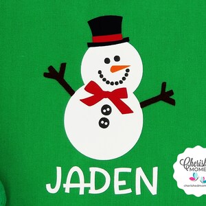 May include: A green background featuring a white snowman design. The snowman wears a black top hat, a red bow tie, and has a carrot nose. The name "JADEN" is printed in white below the snowman. A "Cherished Moments" logo is in the bottom right.