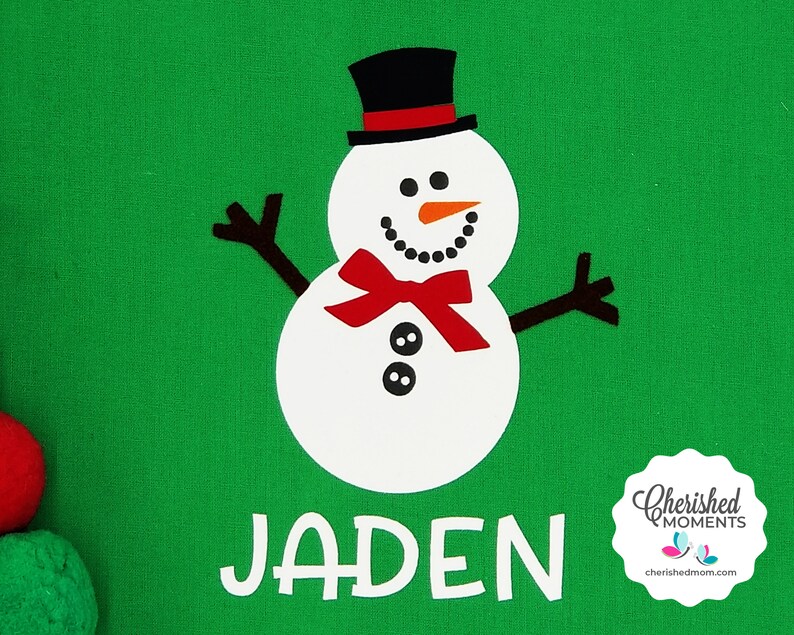 May include: A green background featuring a white snowman design. The snowman wears a black top hat, a red bow tie, and has a carrot nose. The name "JADEN" is printed in white below the snowman. A "Cherished Moments" logo is in the bottom right.