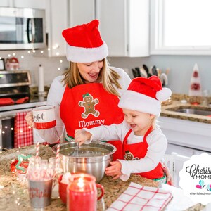 May include: A woman and a child wearing red Santa hats and aprons with gingerbread man designs. The woman holds a mug that says "Joyful." The child stirs a bowl with a whisk. The aprons say "Mommy" and have green trim. Christmas-themed kitchen scene.