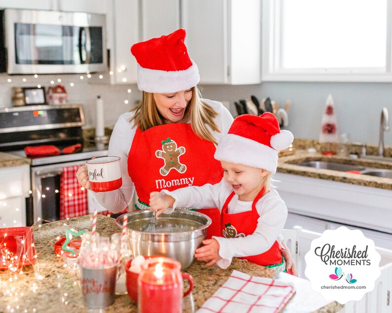 May include: A woman and a child wearing red Santa hats and aprons with gingerbread man designs. The woman holds a mug that says "Joyful." The child stirs a bowl with a whisk. The aprons say "Mommy" and have green trim. Christmas-themed kitchen scene.