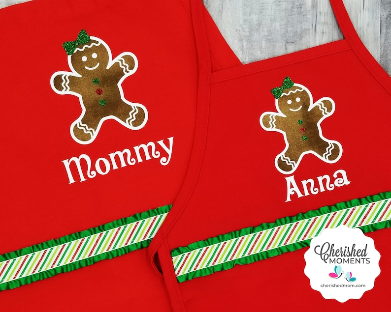 May include: Red aprons featuring gingerbread man designs. One apron says "Mommy" and the other says "Anna". Both aprons have a green and white striped ribbon and a green frill. The aprons are for Christmas.