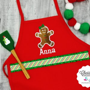 May include: A red apron featuring a gingerbread man design and the name "Anna" in white. The apron has a green and red striped ruffle and a green spatula with a snowman design. A Christmas-themed item.