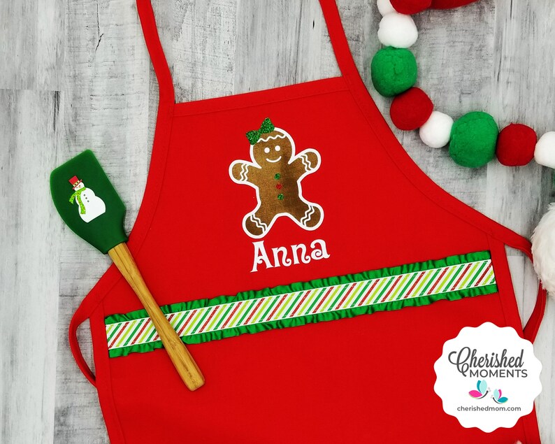 May include: A red apron featuring a gingerbread man design and the name "Anna" in white. The apron has a green and red striped ruffle and a green spatula with a snowman design. A Christmas-themed item.