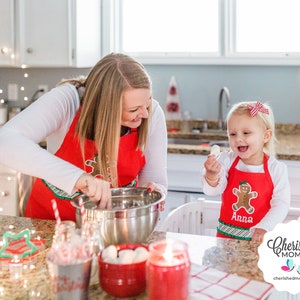 May include: A woman and a young child are in a kitchen, both wearing red aprons with gingerbread man designs. The woman is mixing ingredients in a stainless steel bowl. The child is holding a white object and smiling. Christmas decorations are visible.