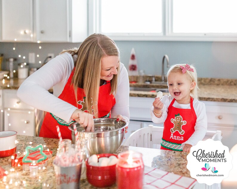 May include: A woman and a young child are in a kitchen, both wearing red aprons with gingerbread man designs. The woman is mixing ingredients in a stainless steel bowl. The child is holding a white object and smiling. Christmas decorations are visible.