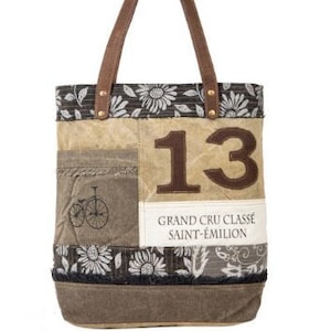 May include: A tote bag featuring a patchwork design with brown and grey tones. The bag displays the number 13, a bicycle graphic, and the text GRAND CRU CLASSE SAINT-ÉMILION. It includes dark brown handles and a floral pattern along the top and bottom.