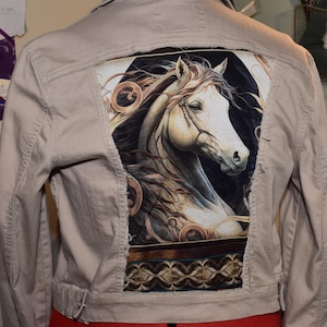 May include: A beige denim jacket with a white horse graphic on the back. The horse is surrounded by gold and brown details. The jacket has a distressed look with frayed edges.