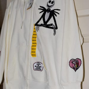 May include: An off-white zip-up hoodie featuring a large black embroidered Jack Skellington character on the chest. A patch with the text Disney The Nightmare Before Christmas is near the pocket, and a heart-shaped character patch is on the sleeve.