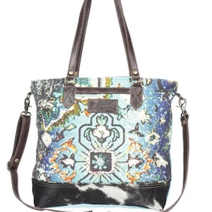 May include: A patterned tote bag with a blue, green, and orange floral design. The bag has brown leather handles and a detachable shoulder strap. The bottom of the bag is black and white cowhide.