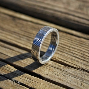 May include: A silver ring with a textured, patterned design. The ring is on a wooden surface.