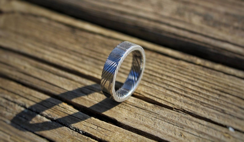 May include: A silver ring with a textured, patterned design. The ring is on a wooden surface.