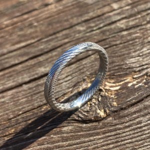 May include: A silver ring with a textured, braided design. The ring is on a wooden surface.
