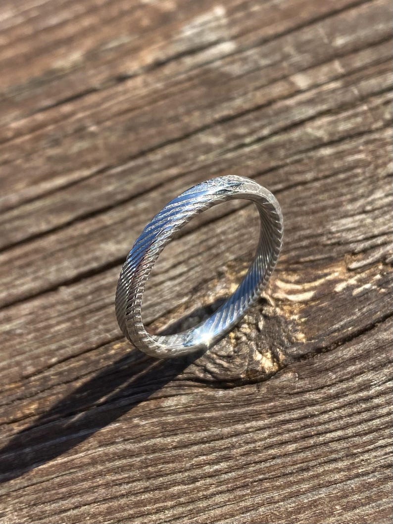 May include: A silver ring with a textured, braided design. The ring is on a wooden surface.